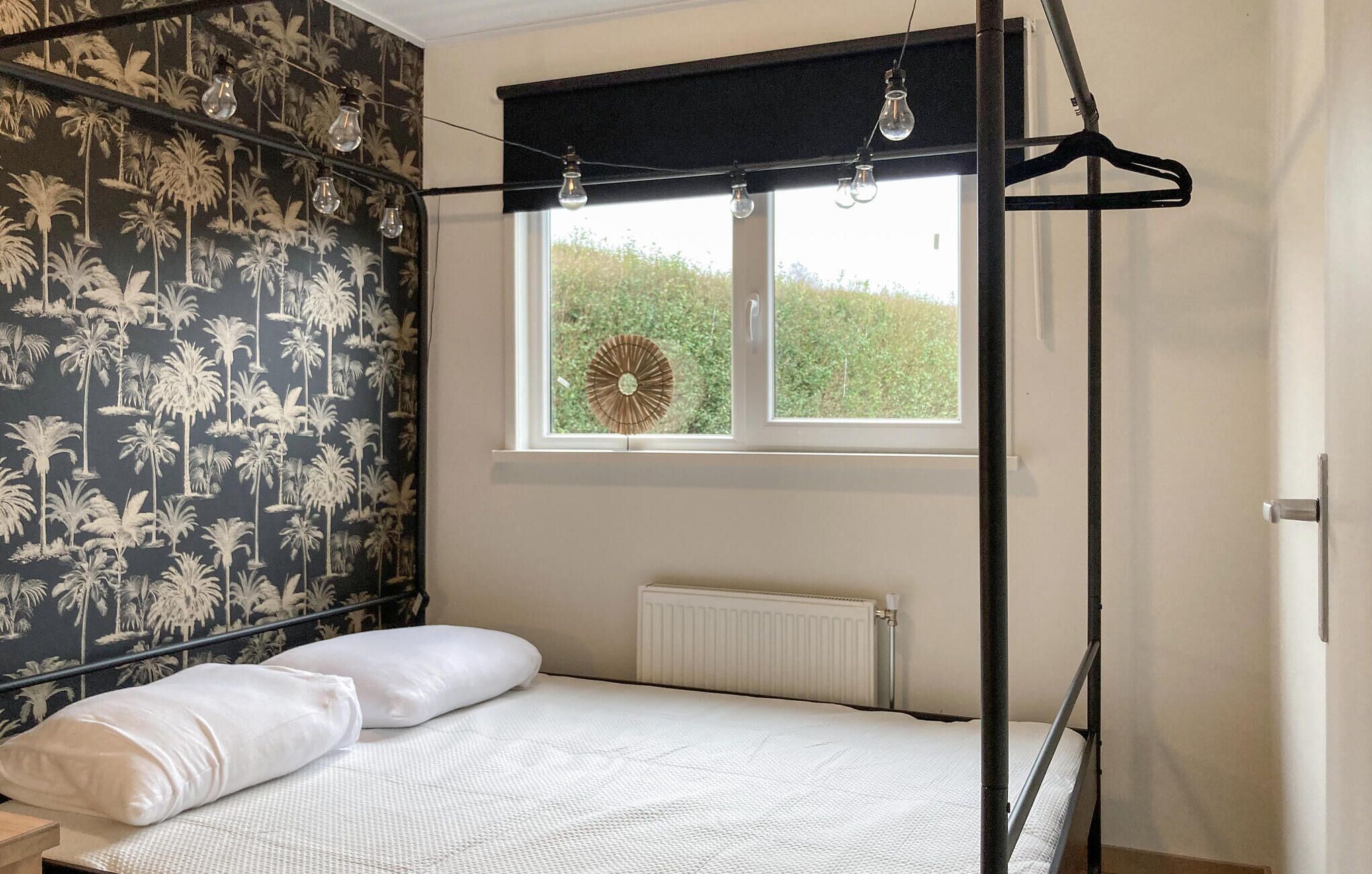 Un dormitorio con una cama, una ventana que ofrece vistas a zonas verdes, un radiador y una pared con papel tapiz de palmeras.