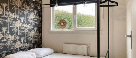 Un dormitorio con una cama, una ventana que ofrece vistas a zonas verdes, un radiador y una pared con papel tapiz de palmeras.