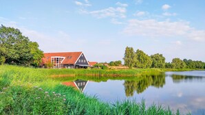 Een traditioneel Nederlands huis met een rood dak, gelegen naast een rustig meer met weelderige begroeiing en bomen op de achtergrond.