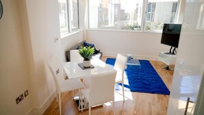 Ein helles, modernes Zimmer mit Esstisch, Stühlen, einem blauen Teppich, einer kleinen Pflanze und einem Fernseher.