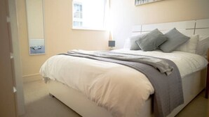Ein ordentlich bezogenes Bett mit grau-weißer Decke, einer Nachttischlampe und einem Fenster mit Jalousien.