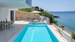 Un área de piscina moderna al aire libre con vista despejada al mar, paredes de piedra y sillones de descanso.