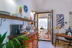 A kitchen with a coffee machine, shelves with dishes, a dining area with chairs, and a table with a lamp. - L'Antro di Dedalo (Sciacca)