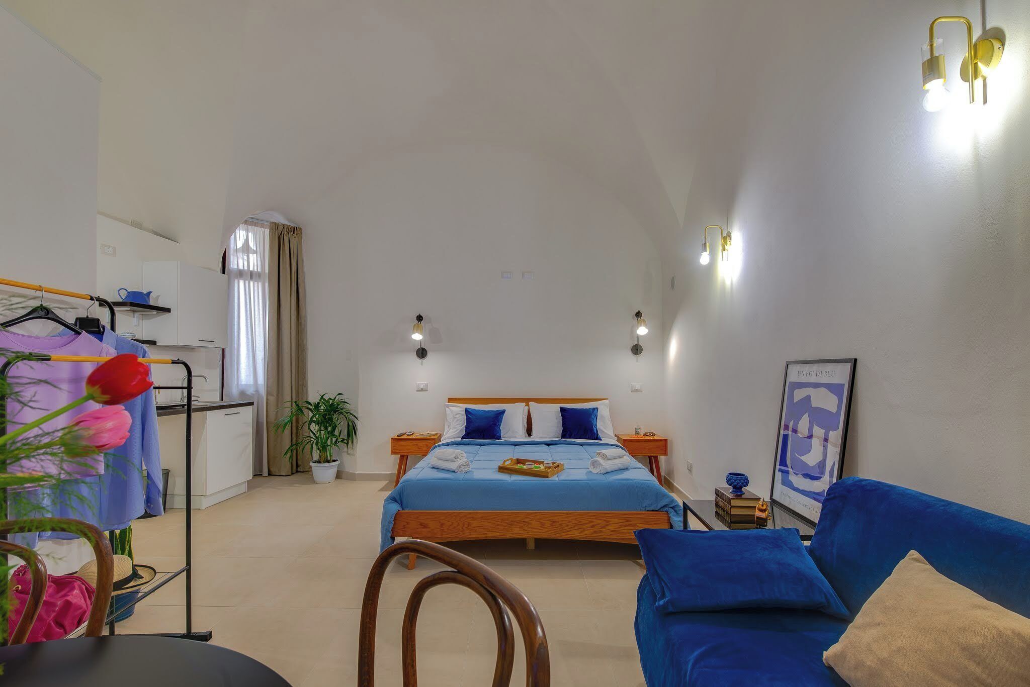 Un dormitorio con cama, sofá azul, escritorio, silla, planta y ventana con cortinas.