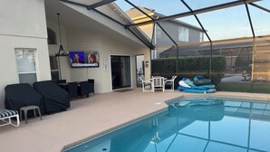 An indoor swimming pool with a covered patio area, seating, and a television screen.