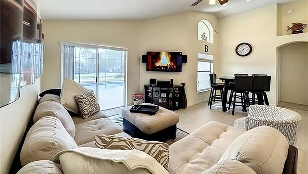 A living room with a sofa, ottoman, and a TV mounted on the wall. There is a ceiling fan, a fireplace, and a dining area with a table and chairs.