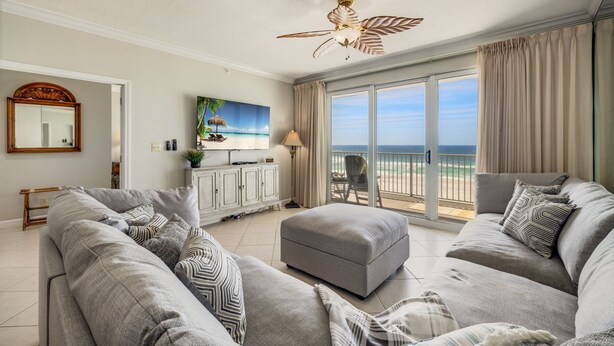 Una sala de estar con un sofá grande de color gris, un reposapiés gris y un ventilador de techo. Hay un ventanal con vistas a la playa.