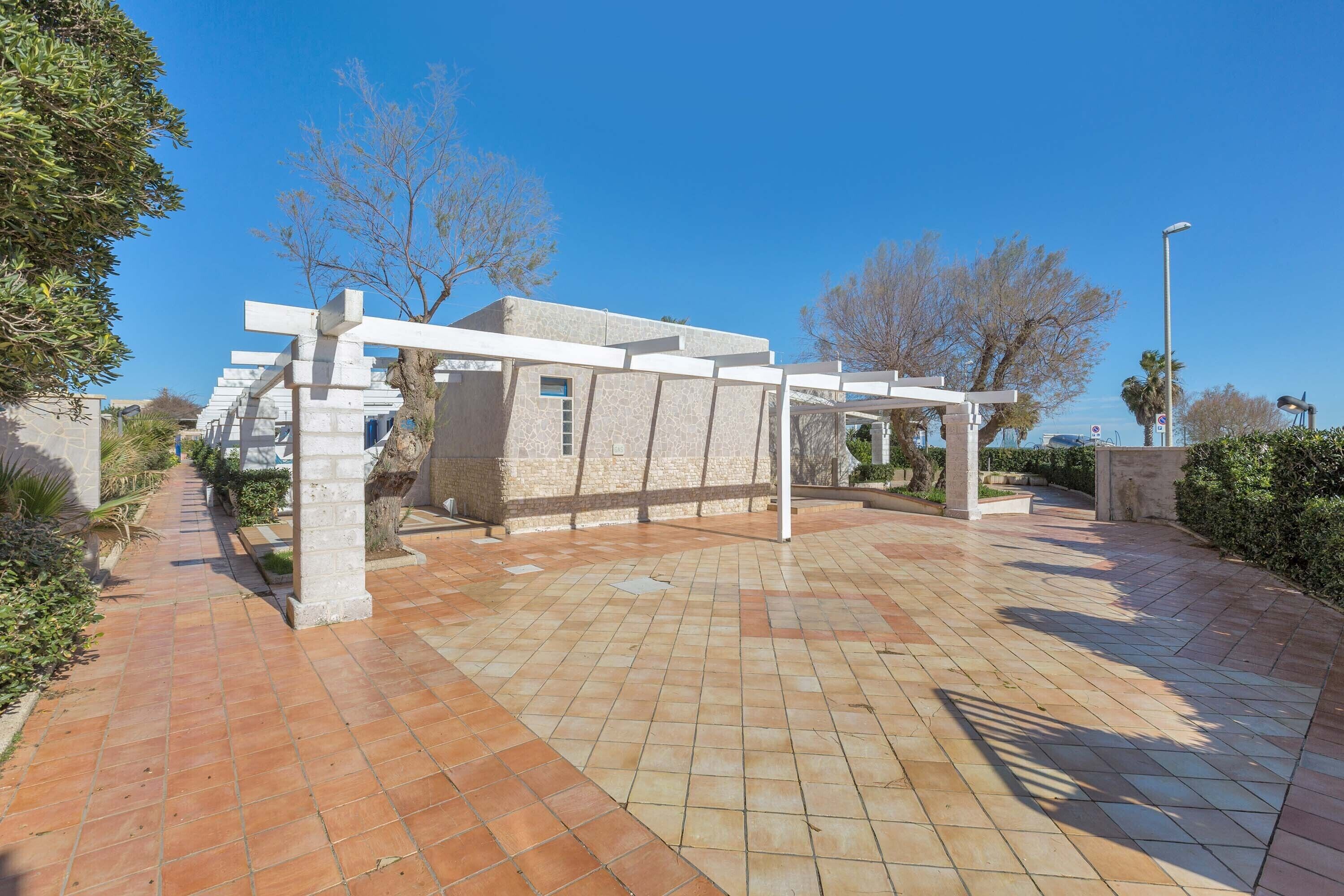 A paved outdoor area with a stone building, white pergola, and a clear blue sky.