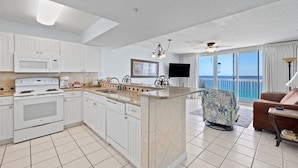 A modern kitchen with white cabinetry, a built-in oven, and a microwave. There's a dining area with a patterned chair and a brown sofa. A large window offers a view of the ocean.