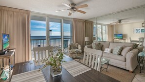 A spacious living room with a balcony overlooking the beach, a ceiling fan, and a television.