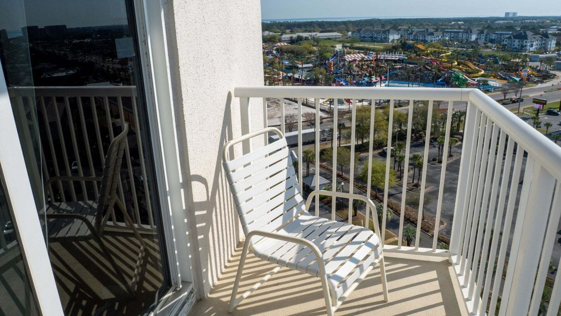 A balcony with a white metal chair and railing, overlooking a cityscape with amusement park attractions.