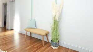 Un pasillo con suelo de madera, un banco, un cojín a rayas y una planta alta en una maceta de fibras.