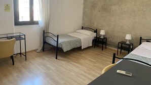 A small, single-bed room with a wooden floor, a desk, two bedside tables with lamps, and a window with curtains.