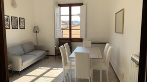 A room with a sofa, dining table, chairs, a floor lamp, and a window with a view of buildings.