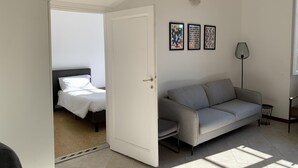 A room with a bed, a grey sofa, and a door leading to another room. There are three framed pictures on the wall.