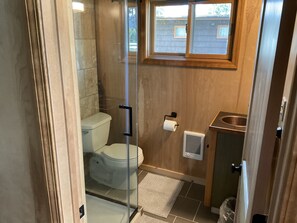 A compact bathroom with a glass-enclosed shower, a toilet, a small sink, and a window above the sink. - Waterfront Cabin in the center of Thompson Falls!! (Thompson Falls)