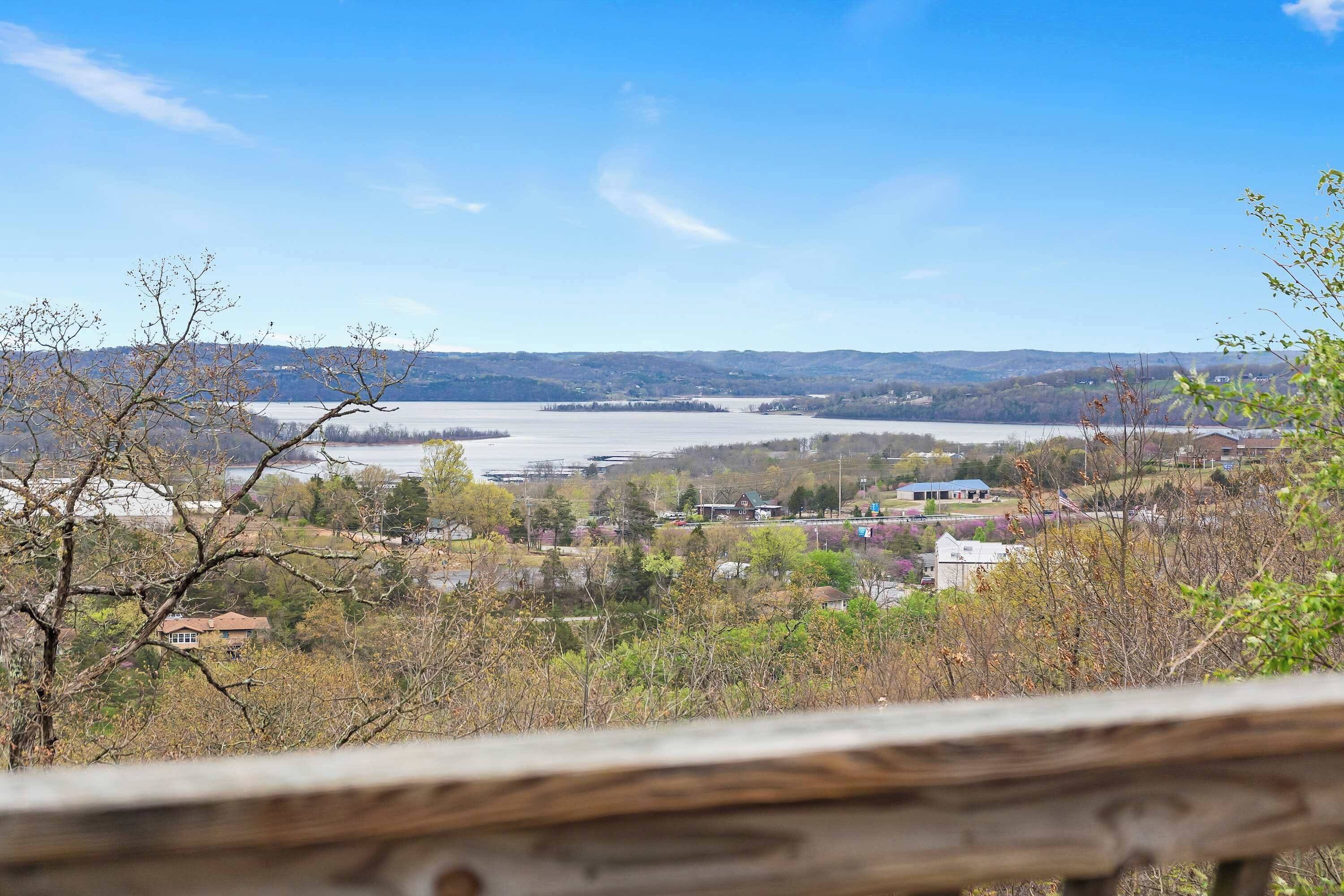 A view from a wooden deck overlooking a lake, with houses and trees in the foreground and hills in the distance.