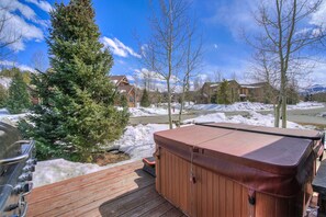 Ein Whirlpool auf einer Holzterrasse mit verschneiter Landschaft und Gebäuden im Hintergrund.