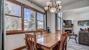 A dining area with a wooden table and chairs, large windows with a view of trees and a house, a chandelier, and a fireplace. - The Oak Lane Chateau: with Private Hot Tub, Close to Golf, Free Ski Shuttle!! (Breckenridge)