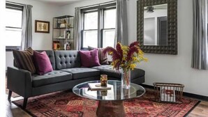 Ein Wohnzimmer mit grauem Sofa, einem runden Couchtisch, einem roten Teppich mit Muster, einem Deckenventilator und einem großen Fenster mit Vorhängen.