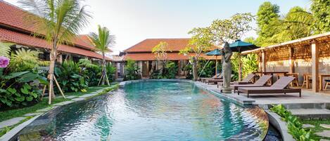 A swimming pool with a circular hot tub, surrounded by lounge chairs and umbrellas, with a tropical garden and a building with a tiled roof in the background.