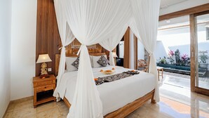 A spacious bedroom with a canopy bed, wooden headboard, bedside table with lamp, and a view of the garden through large windows. - Kudesa Homestay (Sukawati)