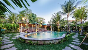 A pool area with lounge chairs and umbrellas, surrounded by palm trees and a stone pathway. - Kudesa Homestay (Sukawati)