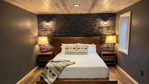 A bedroom with a wooden bed, stone wall, two bedside lamps, and a framed picture on the wall. - Grand Spruce Lodge, Lakefront near Talkeetna & Denali State Park (Trapper Creek)