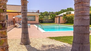 Una piscina rodeada de palmeras, un edificio con techo de tejas y una zona ajardinada.