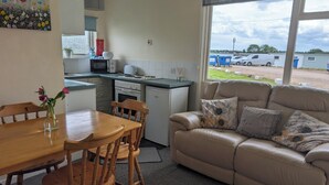 Een knus woongedeelte met een kitchenette, een eethoek met een houten tafel en stoelen, en een beige bank met kussens.