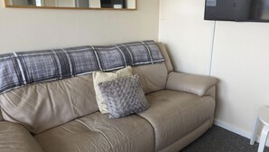Een beige leren bank met een ruitkussen en een pluizig grijs kussen, een klein witte tafel, en een wandgemonteerde flatscreen televisie.