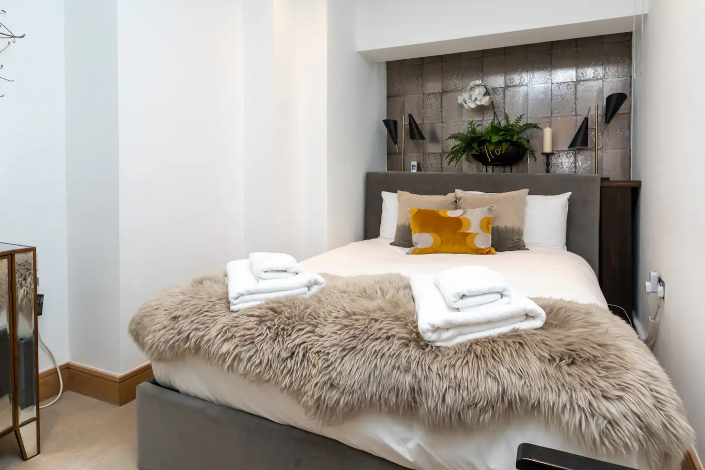 A neatly made bed with a fluffy beige throw blanket, a grey upholstered bench, and a bedside table with a mirror and a lamp.