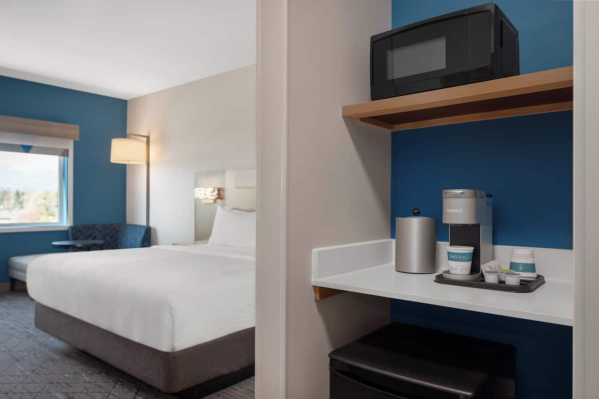 A hotel room with a bed, a desk, a microwave, a coffee maker, and a trash bin.