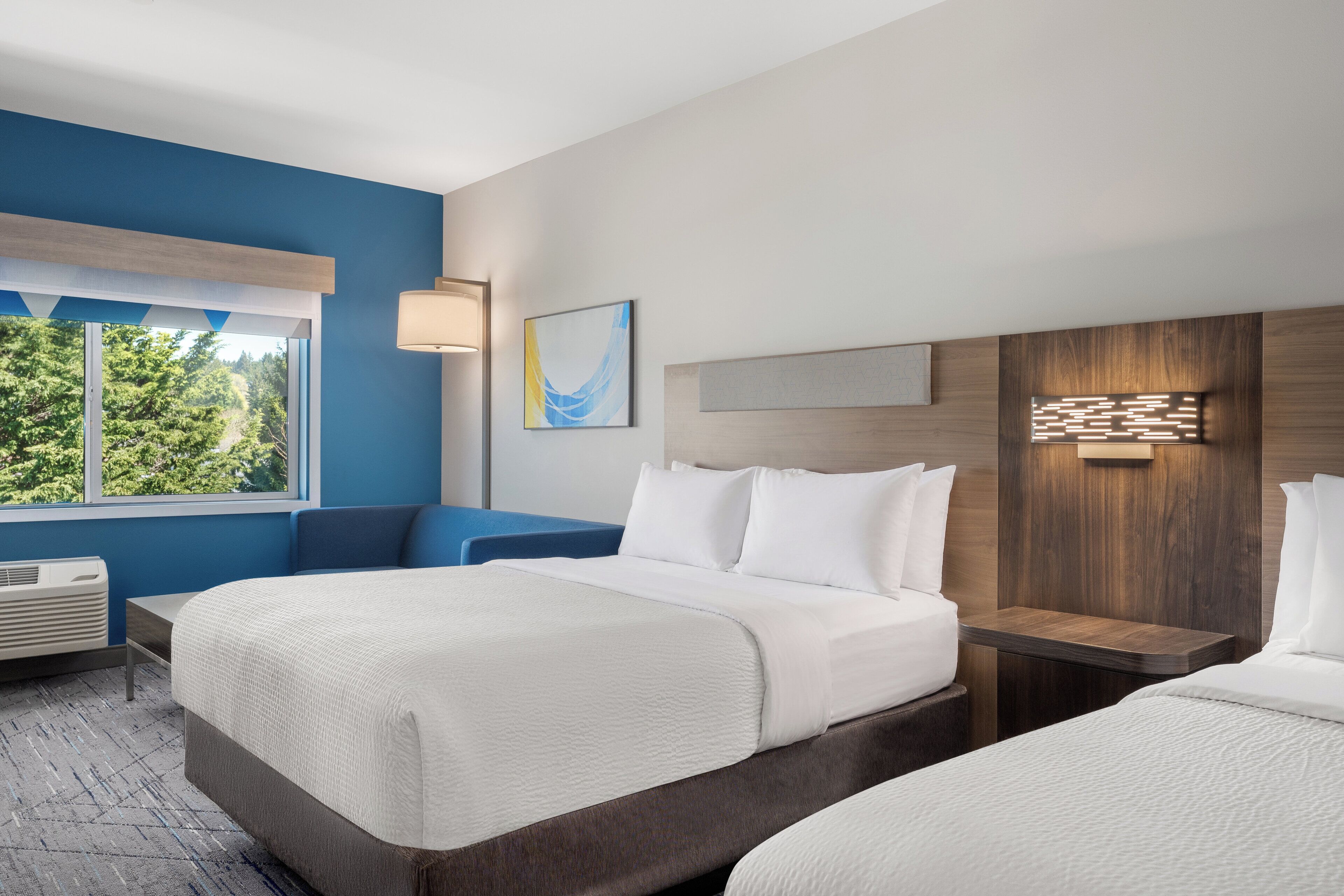 Habitación de hotel con dos camas, un ventanal grande, una lámpara de pared y vistas a zonas verdes.