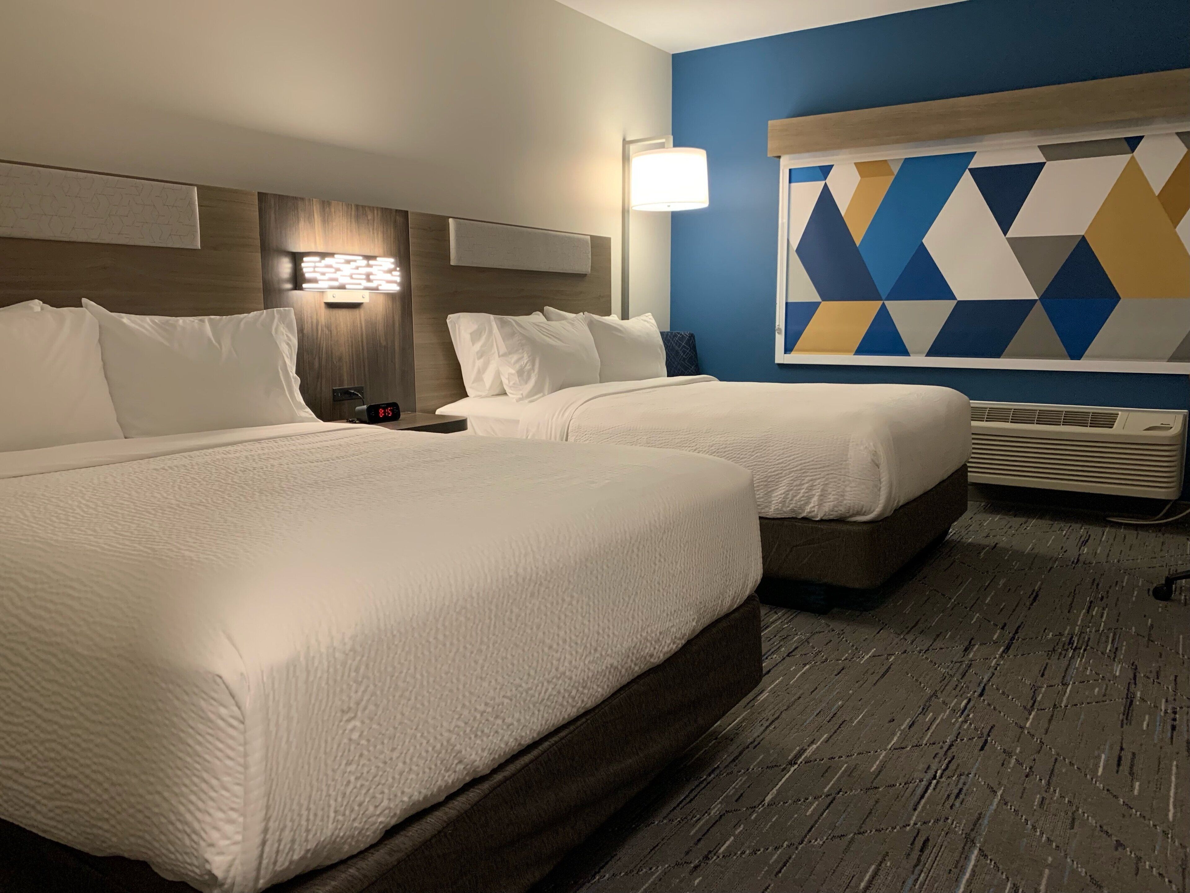 A hotel room with two beds, a wall-mounted lamp, a geometric wall decoration, and an air conditioning unit.
