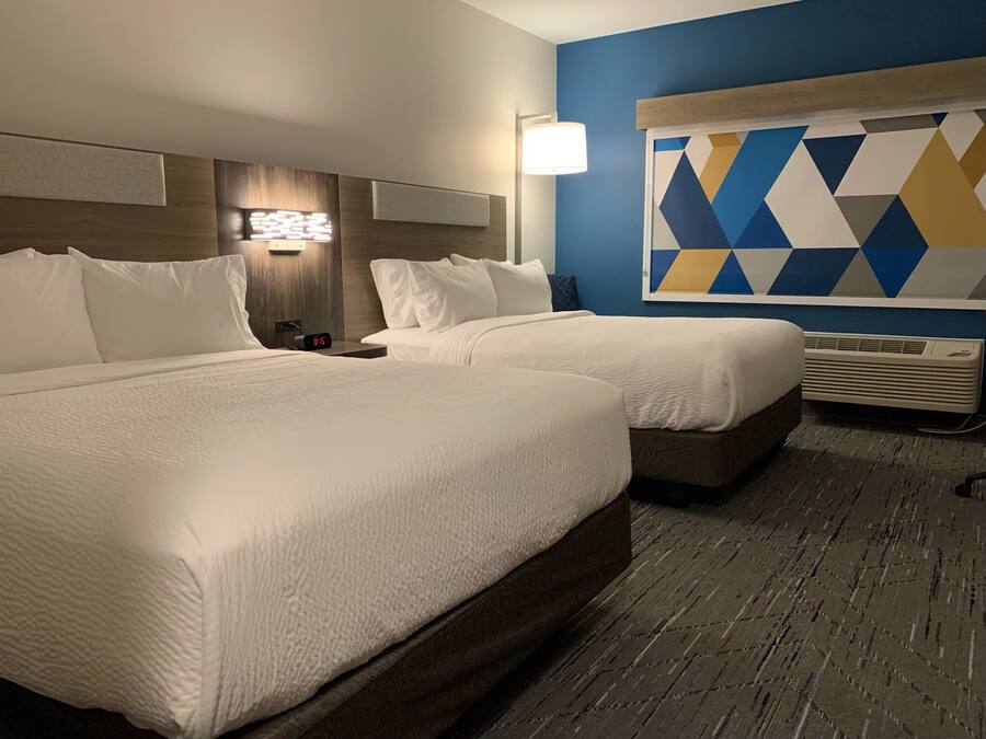 A hotel room with two beds, a wall-mounted lamp, a geometric wall decoration, and an air conditioning unit.