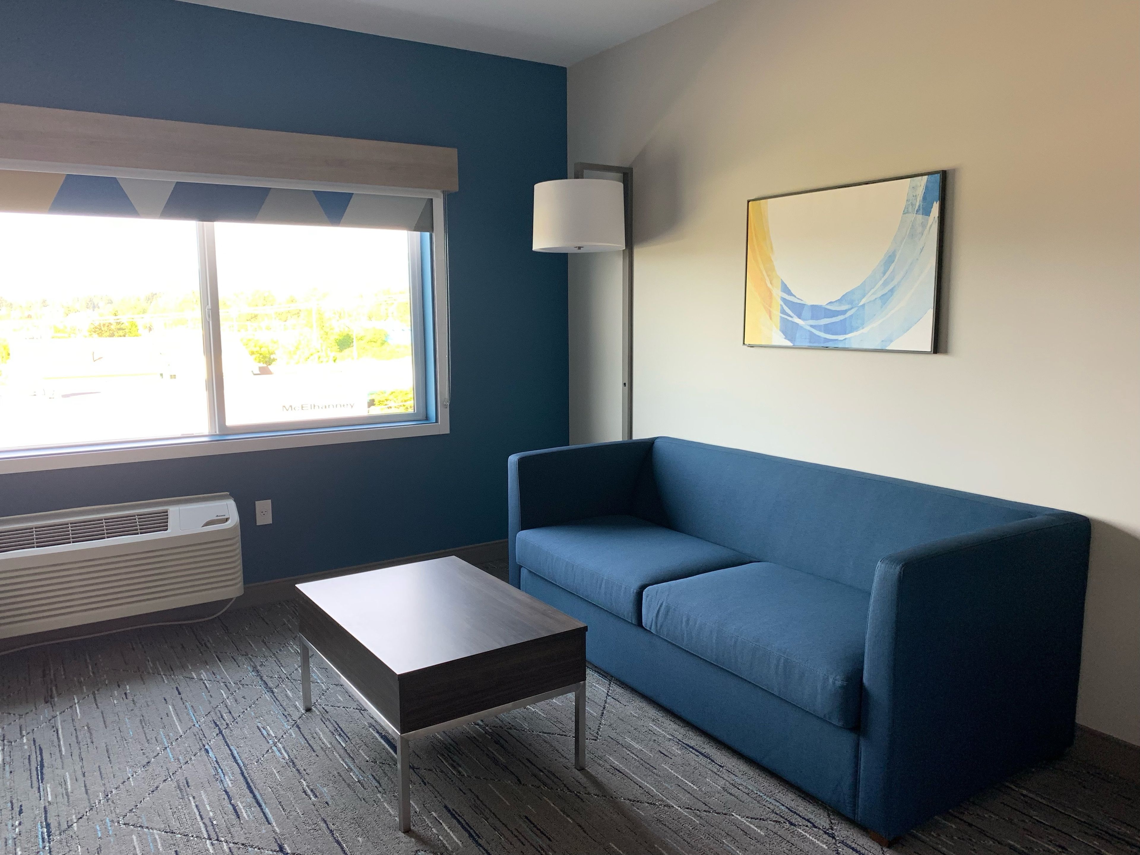 Une pièce avec un canapé bleu, une petite table, un lampadaire et une fenêtre donnant sur un paysage verdoyant.