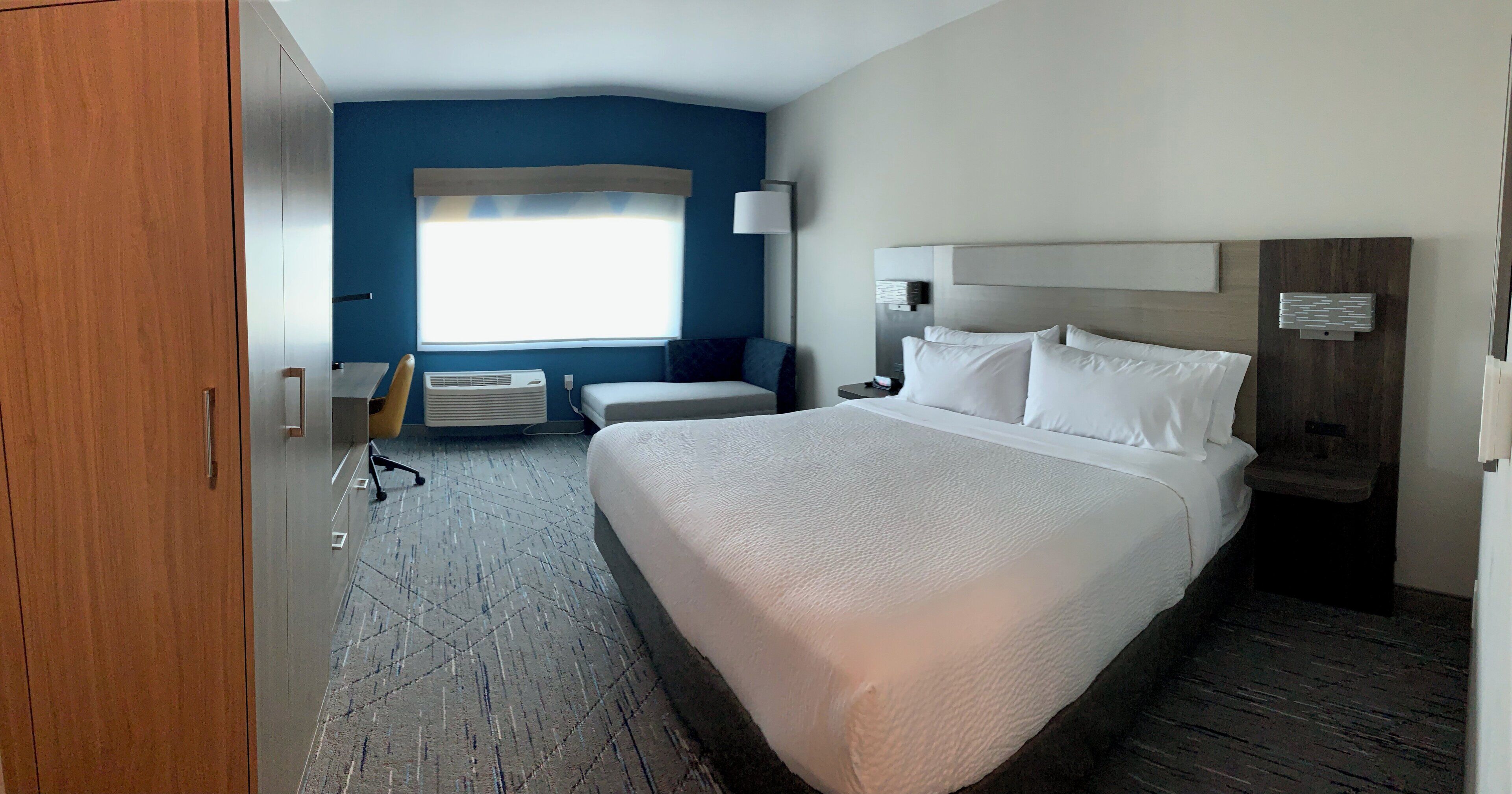 A hotel room with a large bed, a desk, a chair, a window with blinds, an air conditioning unit, and a wooden wardrobe.
