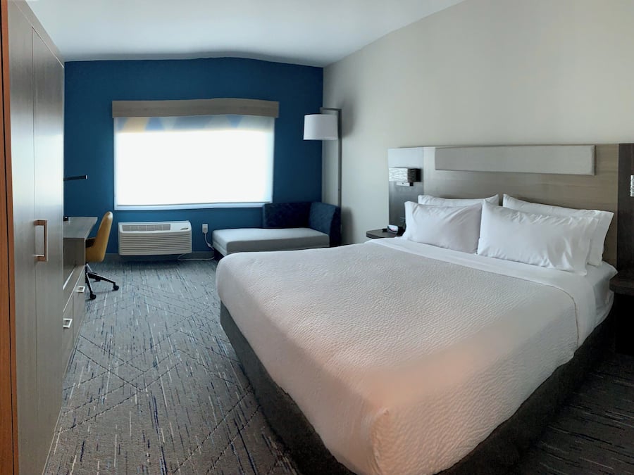 A hotel room with a large bed, a desk, a chair, a window with blinds, an air conditioning unit, and a wooden wardrobe.