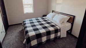 A bedroom with a checkered bedspread, a window with blinds, and a ceiling-mounted smoke detector. - Stockton Lakes’ Rustic Retreat - Cozy & Close (Stockton)
