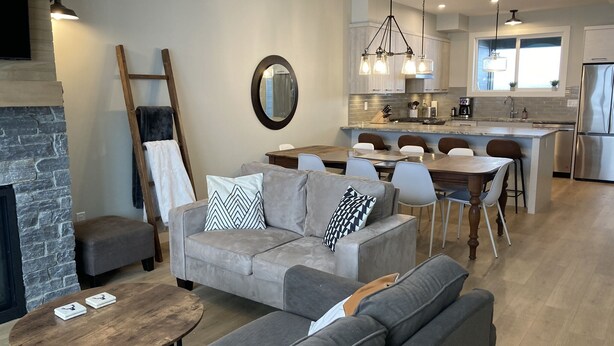 Un salon moderne avec une cheminée, des canapés gris, une table basse en bois et un coin repas avec une cuisine en arrière-plan.