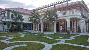 Un edificio grande de dos pisos con techo de tejas rojas, un patio cubierto con palmeras y un jardín ajardinado con senderos decorativos.