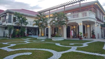 Un edificio grande de dos pisos con techo de tejas rojas, un patio cubierto con palmeras y un jardín ajardinado con senderos decorativos.