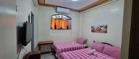 Una habitación con dos camas, televisión, una mesita, ventana con cortinas y un cuadro en la pared.