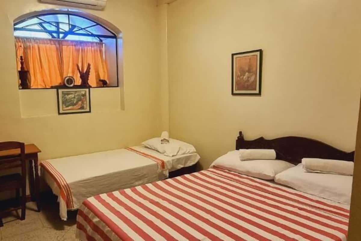 A room with two beds, a desk, a chair, a window with curtains, and a framed picture on the wall.