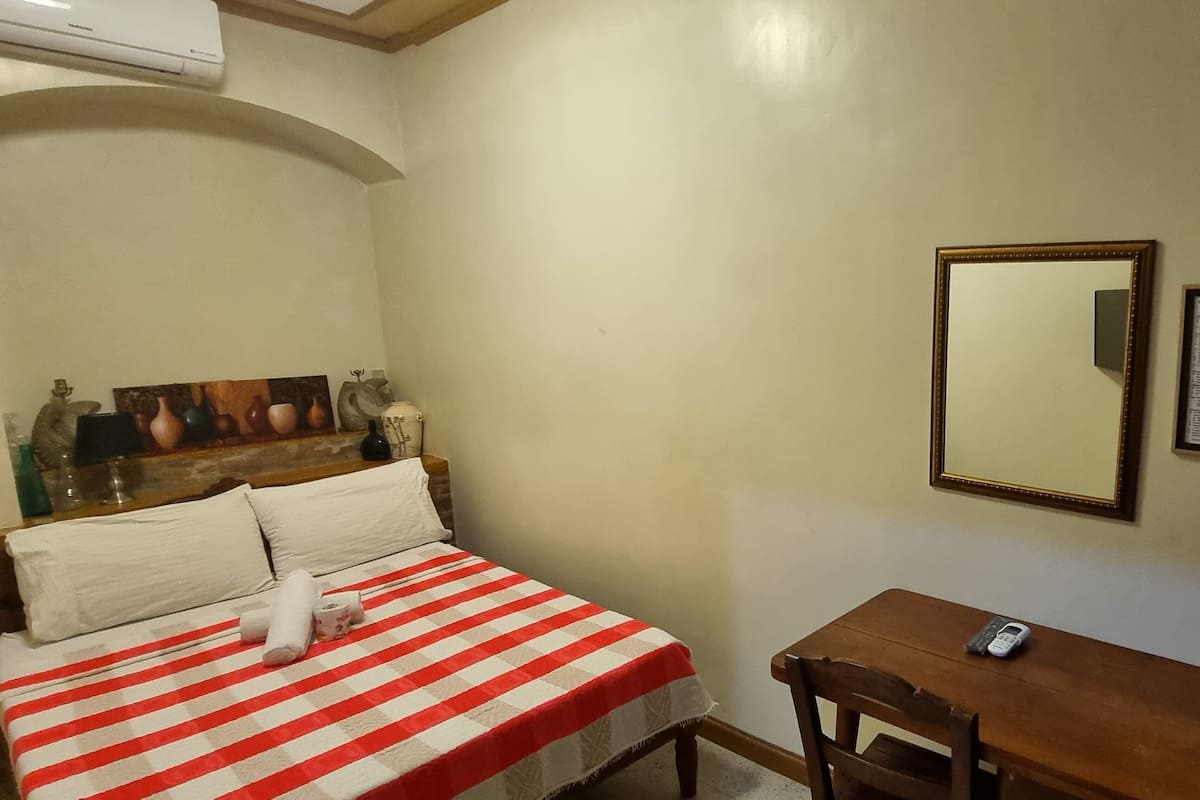 A hotel room with a bed, a small table, a chair, a mirror, and a wall-mounted sign.