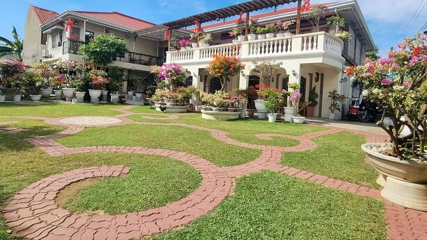 Un edificio de dos pisos con techo de tejas rojas, un balcón con plantas en macetas y un jardín bien cuidado con un camino de ladrillo.