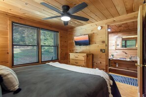 A bedroom with a bed, a ceiling fan, a TV mounted on the wall, and a window with blinds.