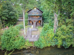 A cabin with a deck, surrounded by dense forest and a body of water.