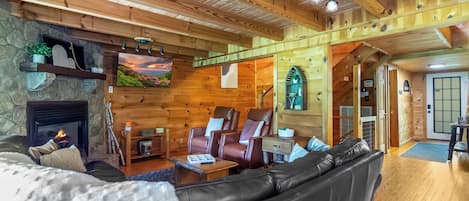 Interior acogedor de una cabaña de madera con chimenea, asientos de cuero, un televisor y una pared de piedra.